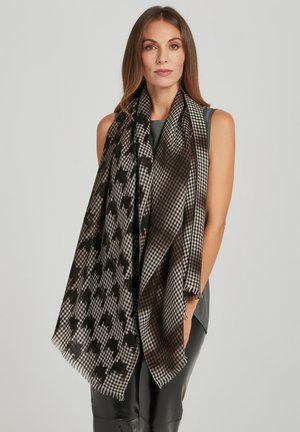 Donna con lunghi capelli castani indossa una grande sciarpa a motivi neri, marroni e beige sopra una canottiera grigia e pantaloni neri.