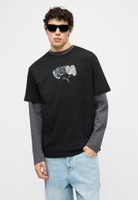 Νέος άνδρας που φοράει μαύρα γυαλιά ηλίου, μαύρο μπλουζάκι με στάμπα πάνω από γκρι μακρυμάνικο, και ανοιχτόχρωμο μπλε τζιν, στέκεται με το ένα χέρι στην τσέπη.