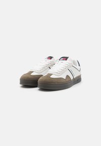 Sneakers med en hvid læderoverdel og brune suede-detaljer, sort og rød branding på tungen, samt en tekstureret grå gummisål.