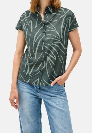 Kvinde med skulderlangt hår har en grøn kortærmet skjorte med bladmønster og lyseblå jeans på, hånden i lommen.