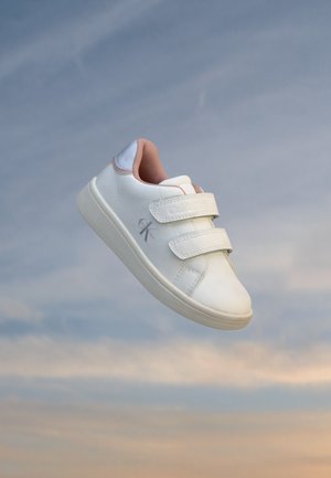 Hvid børnesneaker med to velcro-stropper og lyserød indvendig foring, svævende mod en blød blå og orange himmelbaggrund.