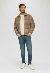 Veste à carreaux brune et grise avec un col, portée sur un pull crème, associée à un jean bleu et des chaussures blanches. Textures lisses et design décontracté.