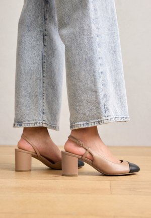 Tacones beige con tira trasera, puntera negra, tacón ancho y correa ajustable. Combinados con vaqueros de pierna ancha y lavado claro.