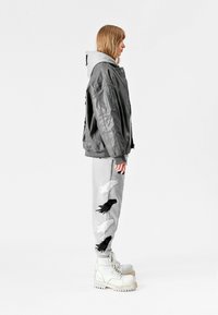 Chaqueta oversized gris sobre una sudadera gris clara, combinada con pantalones de chándal grises con bordado de plumas en blanco y negro, y botas blancas gruesas.