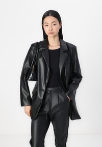 Zwarte faux leren blazer met een notch-kraag, een enkele borstzak en een iets relaxte pasvorm, gecombineerd met bijpassende zwarte broek en een kleine tas.