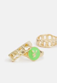 Anéis em tom dourado com uma banda sólida com formas recortadas e um destaque de pedra verde com um motivo de coelho. Textura suave e acabamento brilhante.