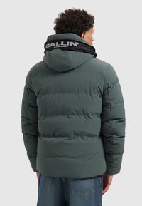 Man met krullend haar, gekleed in een groen gewatteerd jack met opstaande kraag en "BALLIN"-tekst op zwarte trim, gecombineerd met blauwe spijkerbroek, met de rug naar de kijker.