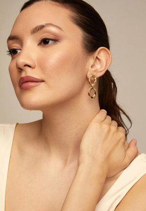 Pendientes de tono dorado con formas ovaladas enlazadas. Acabado metálico liso y pulido. La modelo lleva una camiseta de color neutro y un peinado sencillo.