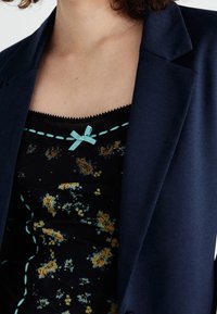 Personne portant un blazer bleu foncé sur un haut noir à motifs floraux jaunes avec un petit nœud bleu près de l'encolure.