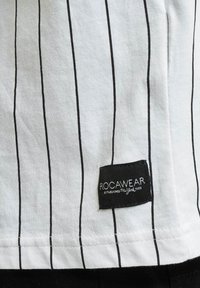 Chemise en coton blanche présentant des rayures verticales noires. Inclut une étiquette noire rectangulaire avec un texte blanc indiquant "ROCAWEAR" en bas.