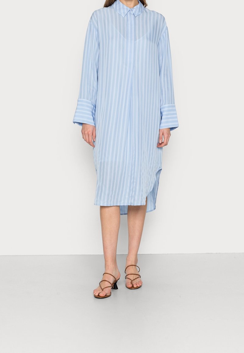 Abito camicia a righe blu chiaro con colletto, maniche lunghe e spacchi laterali, abbinato a sandali marroni con cinturini. Tessuto morbido.