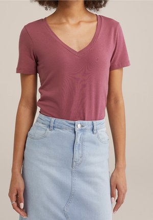 Kvinde iført en kortærmet mauve V-hals skjorte, stoppet ned i et høj taljet lyseblåt denimskørt med frontlommer.