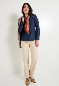 Camisa vaquera de botones, pantalones acampanados color beige y botines marrones. Bufanda con un patrón rojo y naranja atada alrededor del cuello; blazer de cuadros gris sobre el hombro.