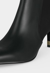 Gros plan d'une bottine en cuir noir avec un bout pointu et un talon fin avec un détail métallique doré.
