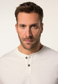 Camicia henley bianca con scollo rotondo, due bottoni neri alla apertura, e un tessuto morbido, indossata da un uomo con la barba curata.