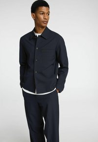 Giacca blu navy realizzata in tessuto texturizzato con colletto a punta, bottoni frontali e taschino sul petto. Abbinata a pantaloni coordinati.