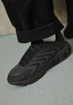 Sneaker sportiva nera con tomaia in mesh e materiale sintetico, lacci neri e suola con ammortizzazione ad aria visibile, indossata con pantaloni neri e calzini bianchi.
