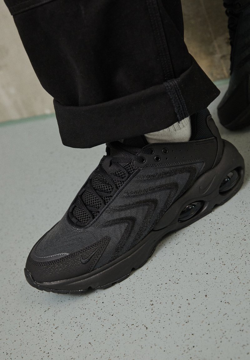 Baskets de sport noires avec une tige en mesh et synthétique, lacets noirs, et semelle visible avec amorti à air, portées avec un pantalon noir et des chaussettes blanches.