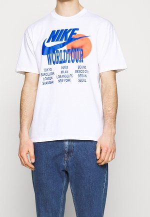 T-shirt graphique blanc arborant "NIKE WORLD TOUR" en bleu avec les noms de villes imprimés en noir. Manches courtes et coupe décontractée.
