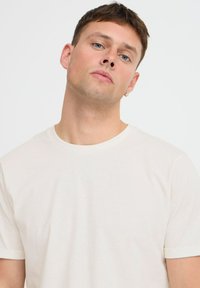 Off-white tričko z měkké, lehké látky; krátké rukávy, kulatý výstřih, jemná textura, přiléhavý střih s ohrnutými okraji rukávů.