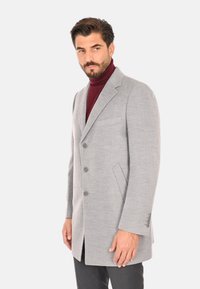 Cappotto grigio in lana con una vestibilità sartoriale, rever a toppa, tre bottoni e tasche laterali. Abbinato a un dolcevita scuro sotto.