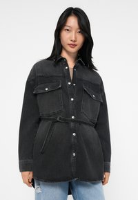 Svart denimskjorta med knappstängning framtill, två stora bröstfickor, bälte i midjan och en något oversized passform.