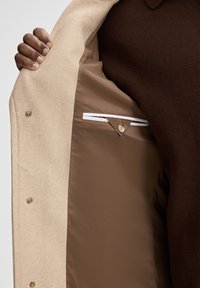 Cappotto in tessuto esterno beige con una fodera marrone, dotato di una tasca marrone e un accento bianco. L'interno ha hardware a pulsante.