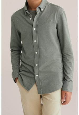 Mann trägt ein olivgrünes Hemd über einem grauen T-Shirt, beige Hose, steht mit einer Hand in der Tasche vor neutralem Hintergrund.