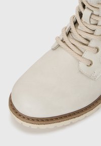 Bottine cheville beige clair en matériau synthétique texturé. Caractéristiques : bout rond, lacets beiges, œillets en métal, et semelle en caoutchouc avec des accents bruns.