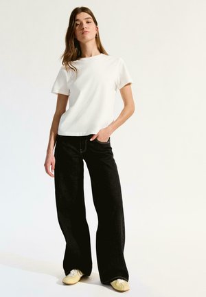 Junge Frau steht entspannt mit einer Hand in der Tasche, trägt ein schlichtes weißes T-Shirt, weite schwarze Jeans und gelbe Turnschuhe vor weißem Hintergrund.