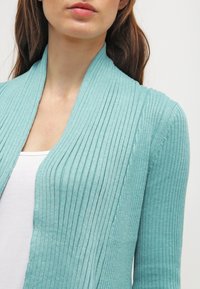 Cardigan de canalé azul claro con un cuello chal, que presenta un patrón de punto texturizado y un ajuste slim, usado sobre una blusa blanca.