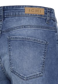 Poche arrière d'un jean en denim bleu avec des coutures visibles et une étiquette de marque en cuir marron clair "ICHI" sur la ceinture.