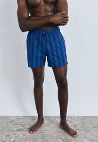 Calções de banho azuis com padrão, cintura elástica e cordão. Apresentam um pequeno emblema com logótipo na lateral. Textura suave, comprimento acima do joelho.