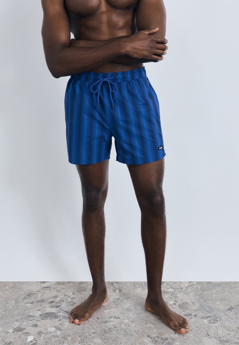 Calções de banho azuis com padrão, cintura elástica e cordão. Apresentam um pequeno emblema com logótipo na lateral. Textura suave, comprimento acima do joelho.