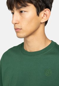 T-shirt in cotone verde con scollo a girocollo e logo ricamato dello stesso colore sul petto, texture morbida.