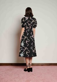 Μαύρο φόρεμα με φουσκωτά μανίκια, διακοσμημένο με πολύχρωμα, αντανακλαστικά σχέδια φιδιού. Μέσο μήκος, συνδυασμένο με μαύρα τακούνια σανδάλια.