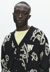 Crni kardigan s kremastim apstraktnim uzorkom, s jednim logotipom na lijevoj strani prsa i gumbima sprijeda, nošen preko svijetlo žute košulje.