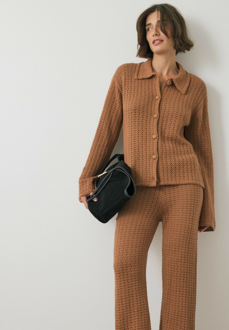 Knitted brown cardigan with a collar and buttons, paired with matching textured trousers. Model holds a black handbag with a gold accent.