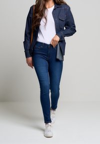 Chaqueta de mezclilla con diseño de botones, combinada con una camiseta blanca y unos pantalones ajustados de mezclilla azul oscuro, complementada con zapatillas blancas.