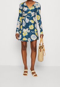 Robe portefeuille florale à manches longues, dans des tons de bleu et de jaune, associée à un sac à main tressé et à des sandales à enfiler beiges.