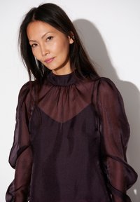 Paarse doorschijnende blouse met opgeblazen lange mouwen, hoge hals en geplooide details, met een subtiele glans en gelaagd ontwerp.