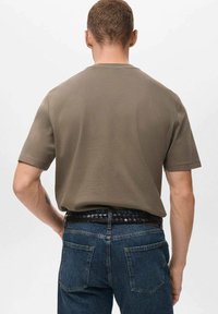 Mand vist fra bagfra iført en olivengrøn kortærmet skjorte, som er stoppet ind i et par blå jeans med et sort vævet bælte.