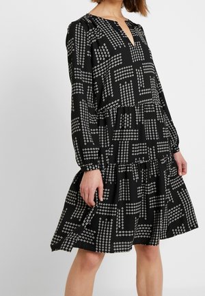 Frau trägt ein locker sitzendes schwarzes Kleid mit langen Ärmeln und einem weißen geometrischen Muster, steht vor einem schlichten hellen Hintergrund.