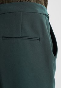 Ciemnozielone spodnie o gładkiej fakturze, z tylną kieszenią z poziomym szwem i szlufkami na pasek, zaprojektowane w klasycznym stylu.