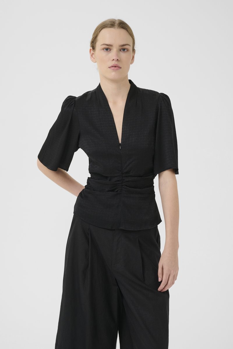 Black short-sleeve top with a deep V-neck, subtle texture, and gathered detail at the waist. Paired with wide black trousers.