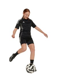 Jersey y pantalones cortos deportivos negros con rayas blancas, jugador de fútbol en acción, equilibrándose sobre un balón de fútbol con un patrón blanco y negro.