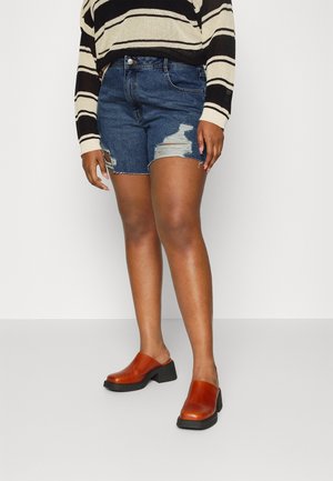 Persoon met een zwart-beige gestreepte trui, distressed blauwe denim short en bruine muiltjes met hak staat tegen een effen witte achtergrond.