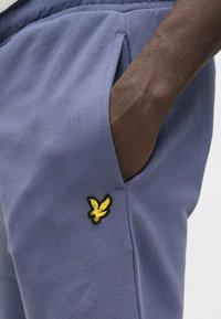 Lyseblå bomuldsjoggers med sidelommer, med en sort og gul ørn-logo broderet på venstre side. Glat tekstur.