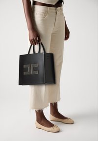 Black leather bag with a structured design, double handles, and a prominent logo detail in gold. Worn with beige, cropped pants and flats.
