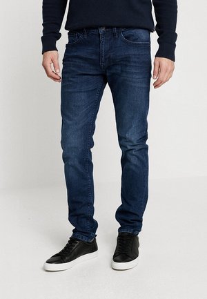 Homme portant un jean slim bleu foncé, un pull noir et des baskets noires à semelles blanches, debout sur un sol clair uni.